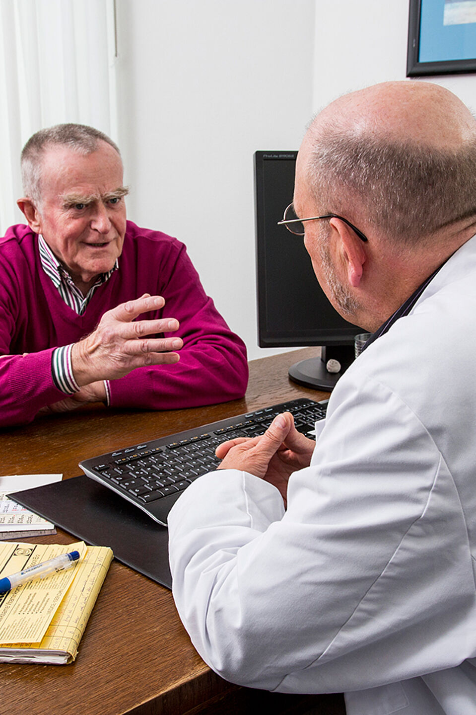 Patientengespräch Ein Arzt am Schreibtisch ist von schräg hinten zu sehen, wie er mit einem Patienten spricht.