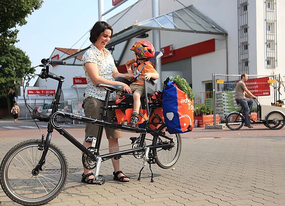 Kindertransport im Kindersitz hinten Frau schnallt Kind im Kindersitz an. Es sitzt hinten auf dem Fahrrad, dahinter sind noch Gepäcktaschen angehängt.