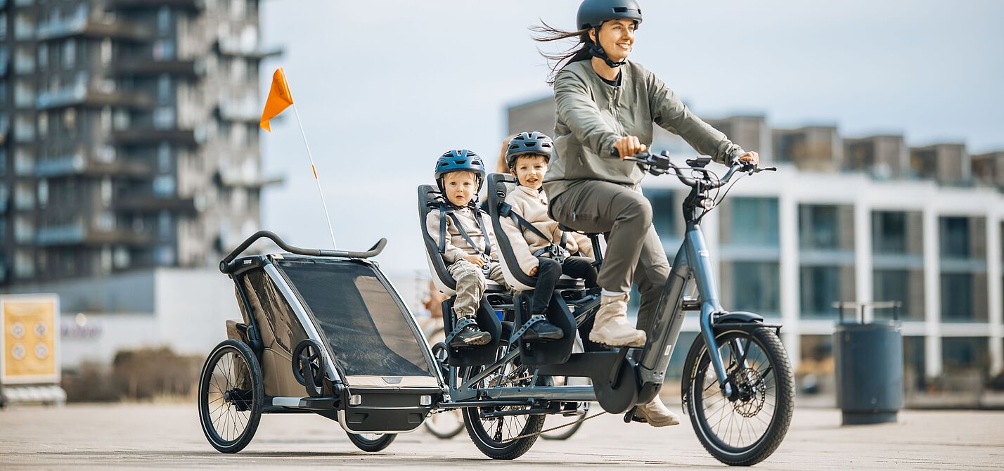 Frau fährt ein Lastenrad Longtail, auf dem langen Gepäckträger hinten sitzen zwei Kinder in je einem Kindersitz. Das Fahrrad zieht zudem einen Kinderfahrradanhänger.